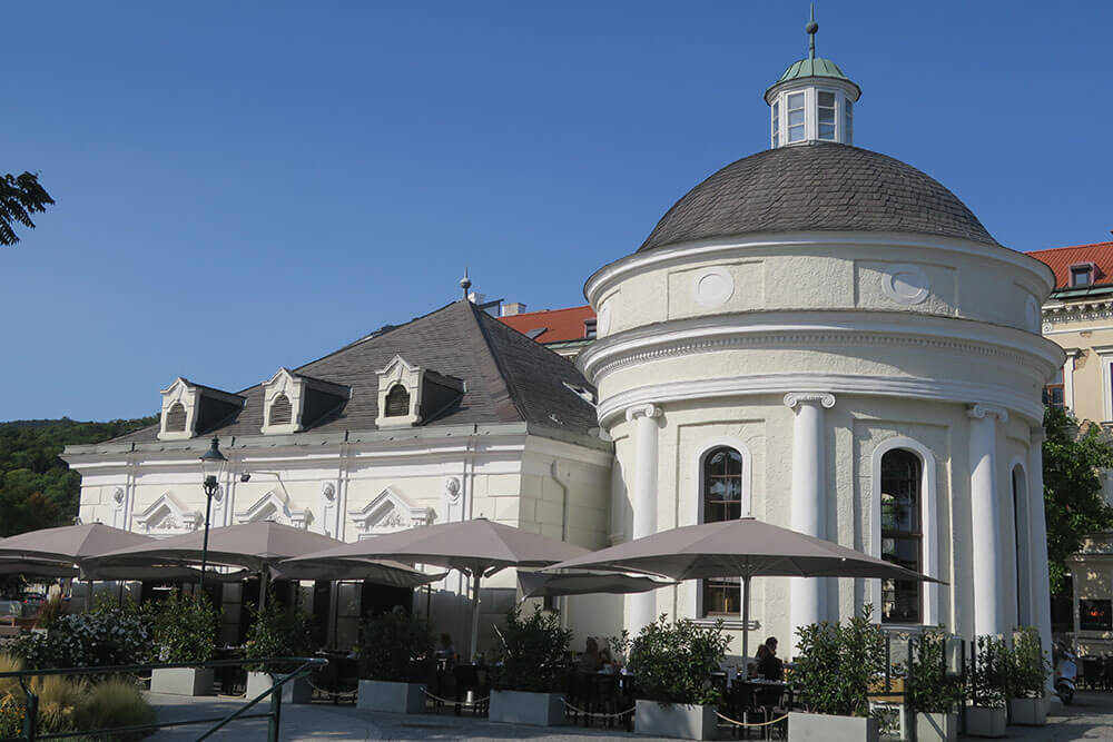 Josefsbad. Helle Fassade - eckiges Gebäude und rundes Gebäude mit Kuppeldach. Schirme auf der Terrasse im Vordergrund.