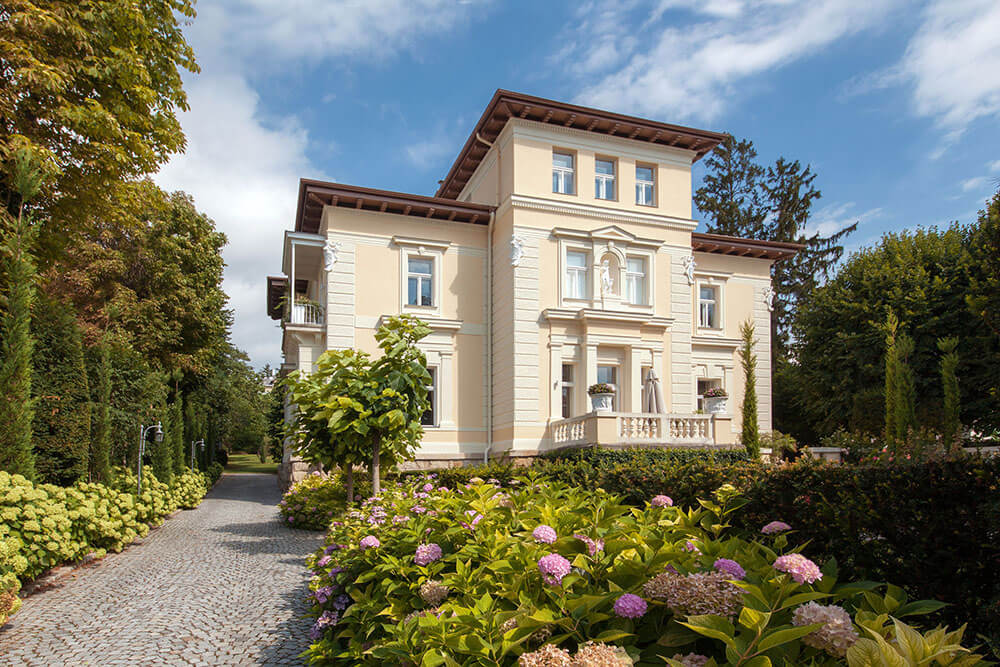 Dreistöckige Villa im Adria-Stil, helle Fassade mit Palladio-Motiv; im Vordergrund Garten mit Blumen, links Bäume und ein Weg.