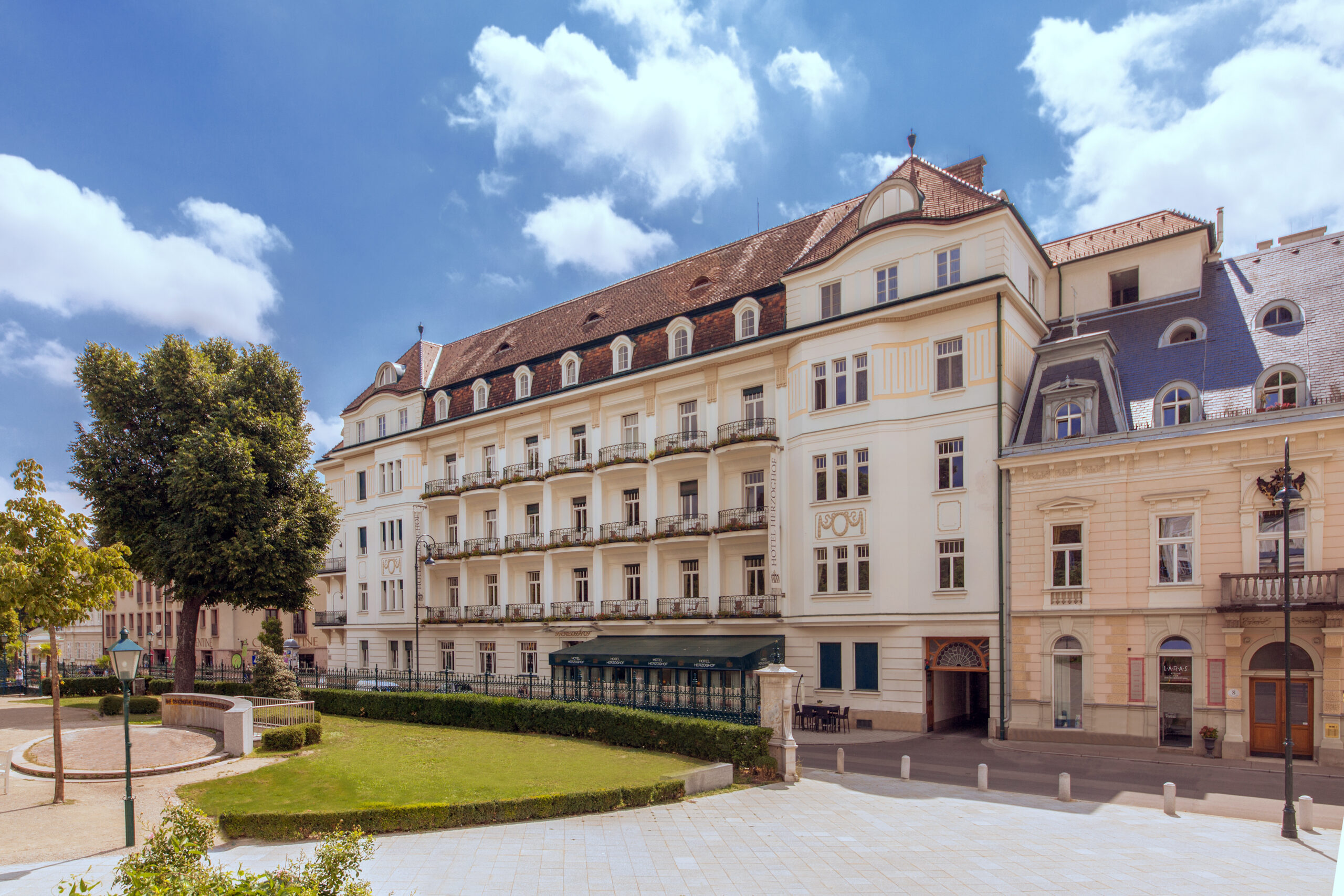 Viergeschossiges Gebäude mit Balkonen, Mansarddach und heller Fassade an einer Straße, links Grünfläche.