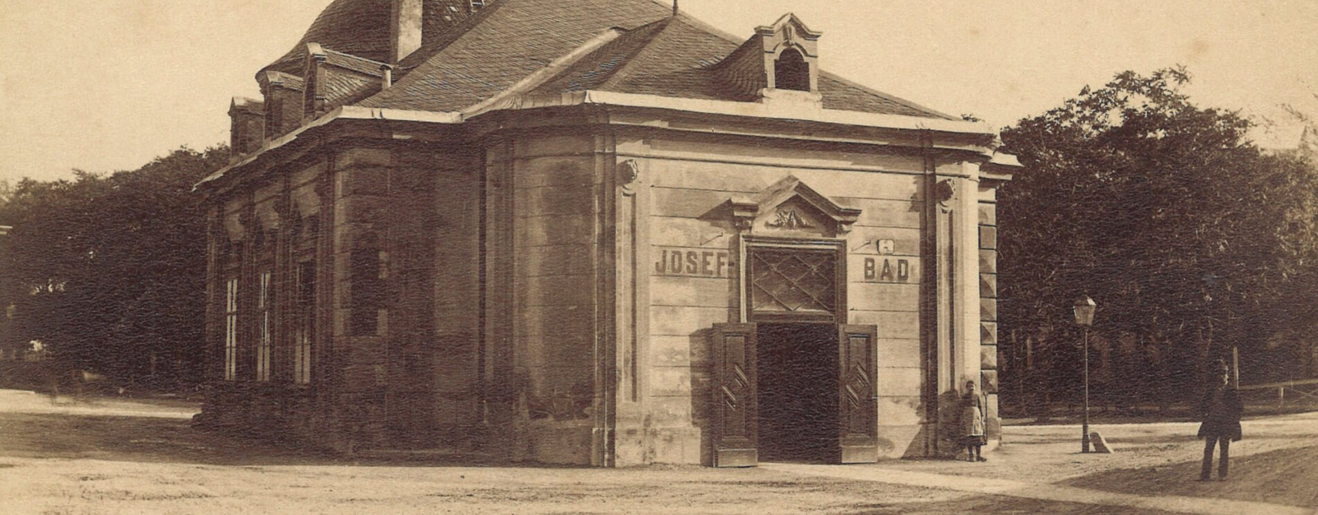Blick auf den Eingang: freistehendes Badehaus aus Stein, davor unplanierte Straße mit Sand und Erde.