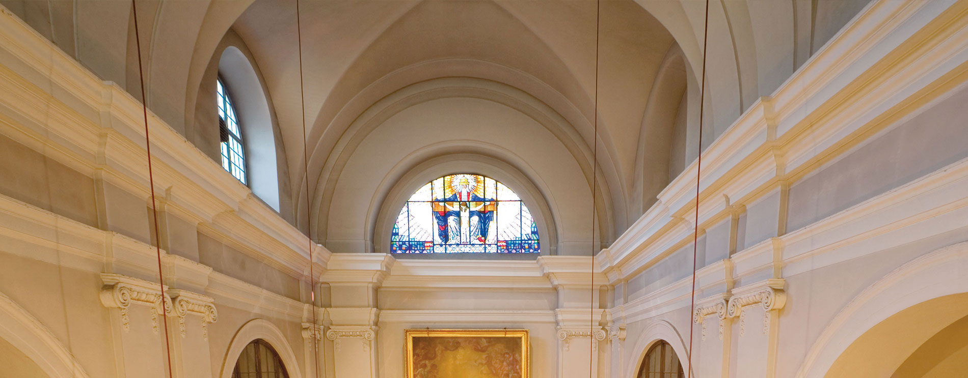Halbrundes Oberlicht mit buntem Glas mittig über dem Altar; darunter Gesims und Kapitelle der Wandsäulen.