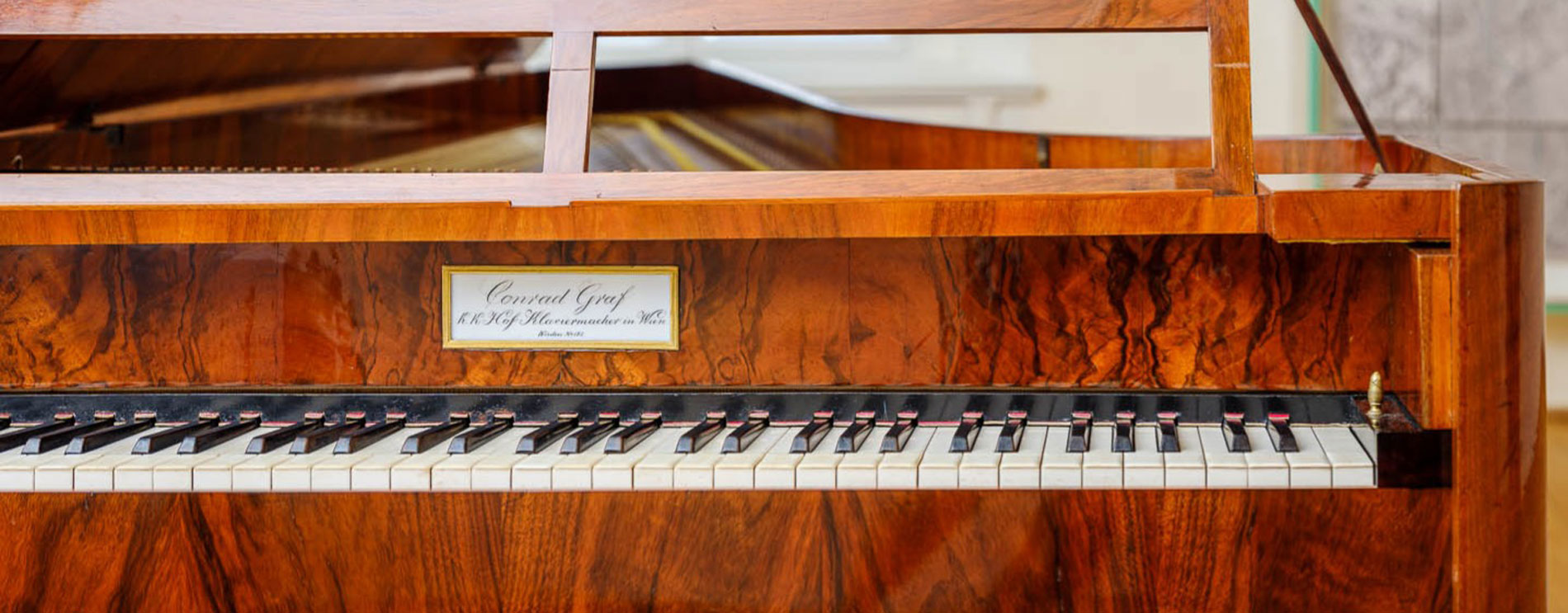 Tastatur eines Klaviers mit heller Holzmaserung; mittig ein kleines Schild mit dem Namen „Conrad Graf“.