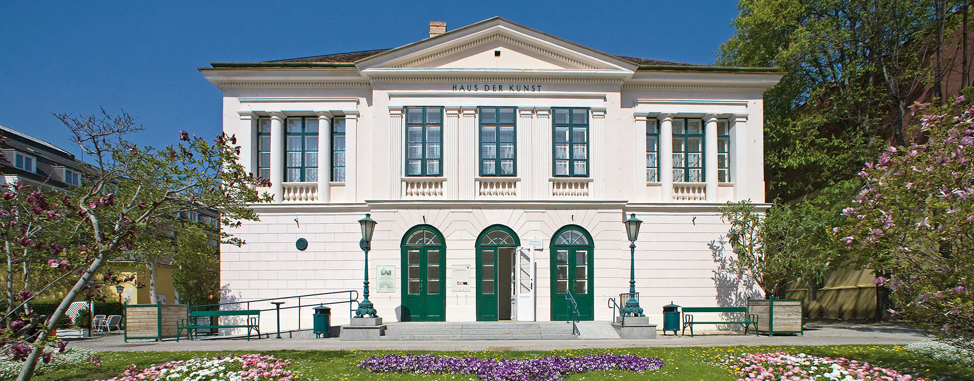 Weißes zweistöckiges Gebäude mit grünen Kastenfenstern und Mittelgiebel; davor malerisch bunte Blumen.