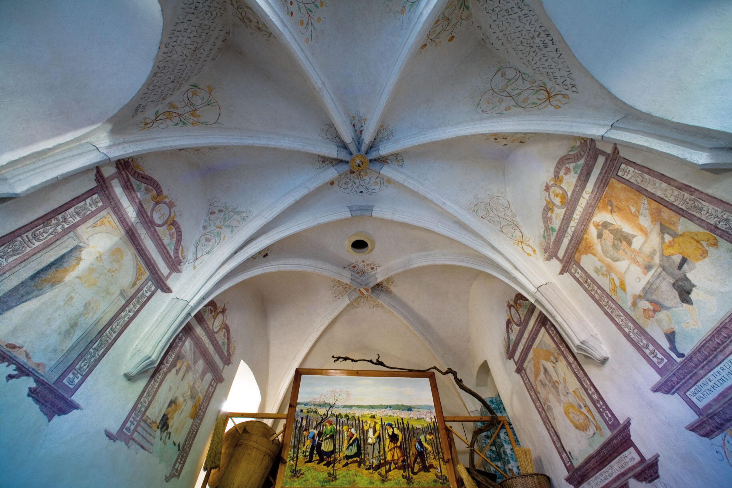 Blick auf die weiße Kapellendecke mit gotischem Kreuzrippengewölbe; christliche Wandmalereien, dazwischen Weinstock und Werkzeug zur Weinlese.