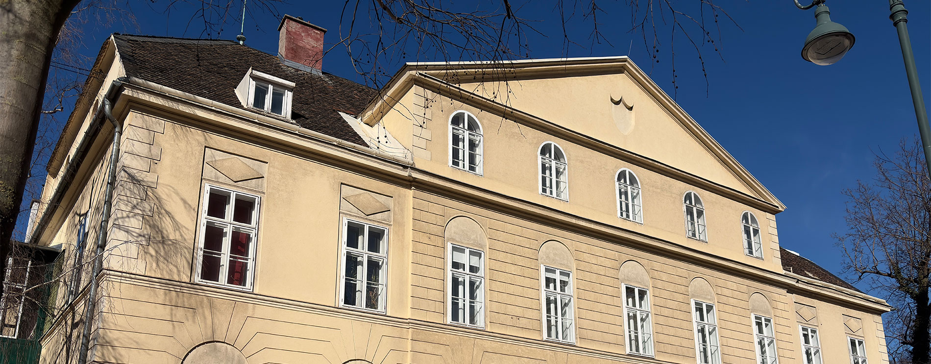 Symmetrischer zweigeschossiger Bau mit drittem Stock unter einem mittigen Giebel mit Rundbogenfenstern; davor Bäume und Straße.