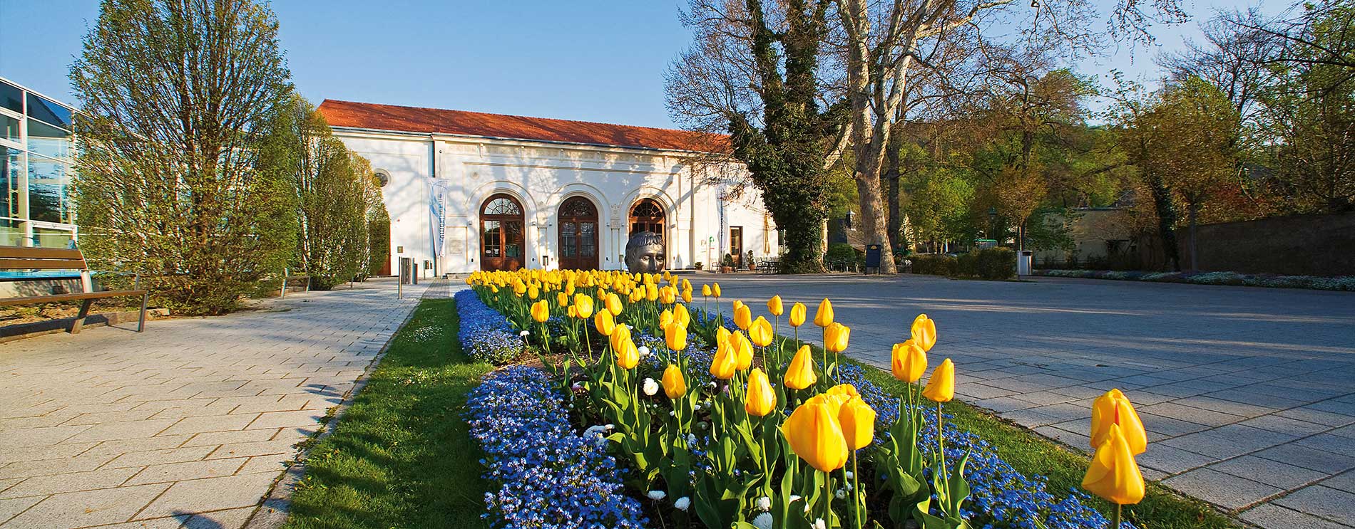 Blick auf den heutigen Eingang mit steinernen Torbögen; davor ein Tulpenbeet, links die Glasfassade der Römertherme.
