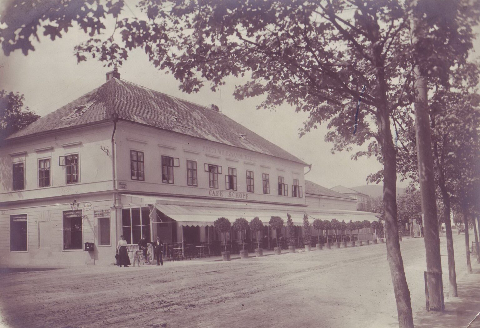Zweigeschossiges Haus mit Walmdach, Café mit Markise und Sitzplätzen entlang der Straße; Schwarzweißfoto.