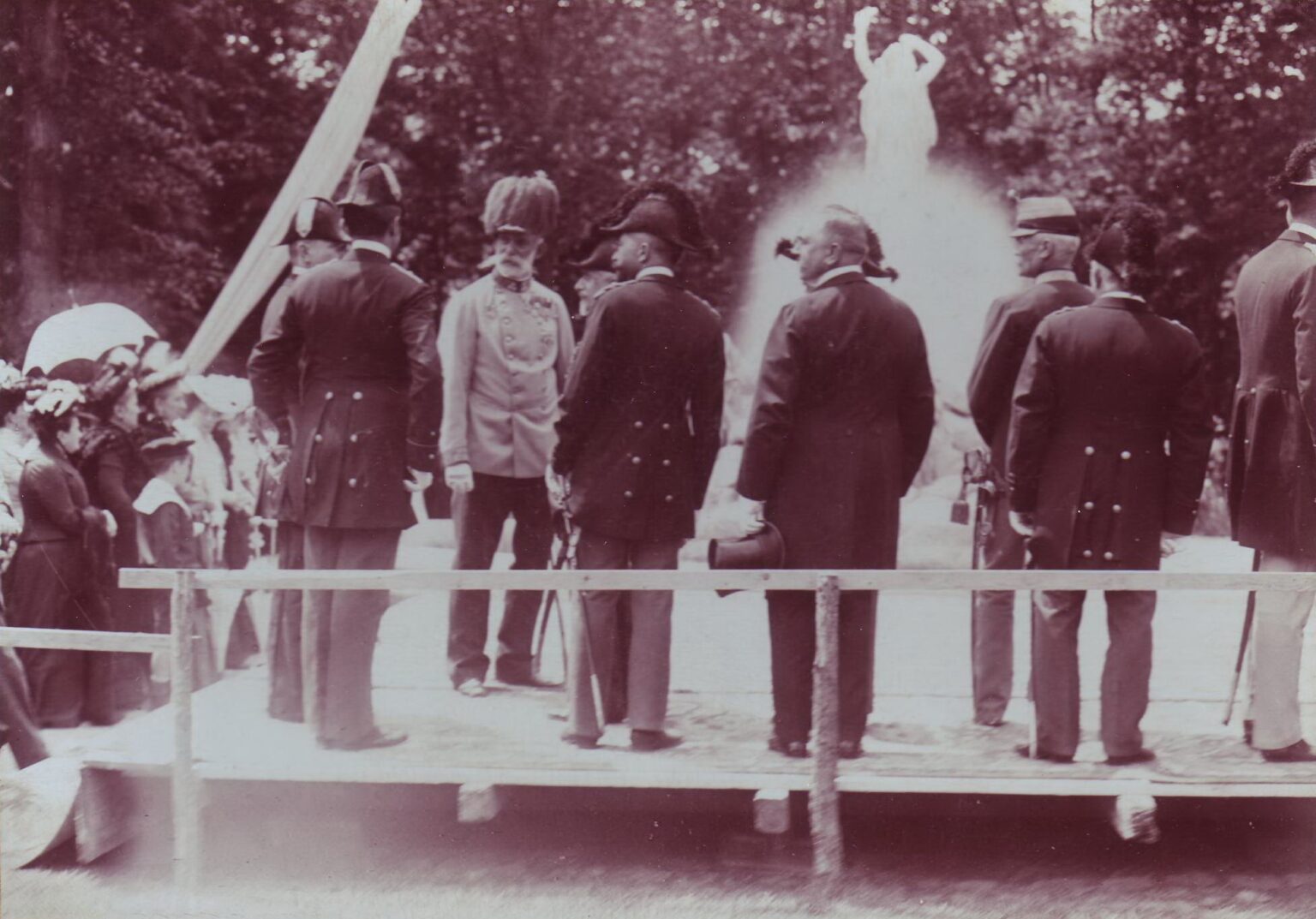 Mehrere Männer in Uniform oder Anzug auf Holzbühne vor dem Undinebrunnen mit Sprühregen im Hintergrund; Schwarzweißfoto.
