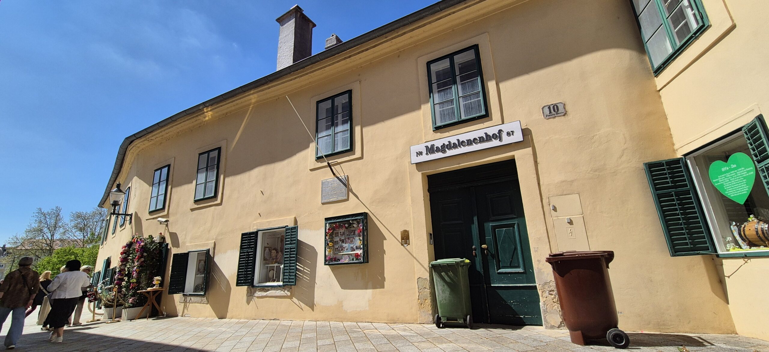 Zweigeschossige Fassade mit grünen Fenstern, im Erdgeschoss Fensterläden, strahlend blauer Himmel.