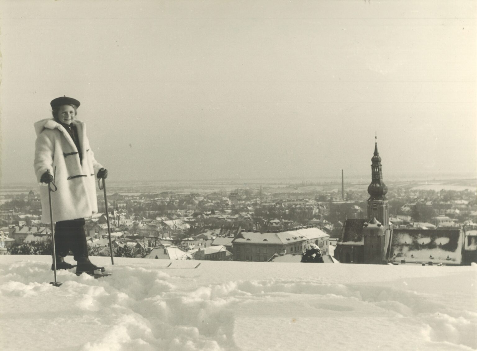 Schifahrerin posiert vor verschneiter Stadtansicht; links hinten die Pfarrkirche; Schwarzweißfoto.