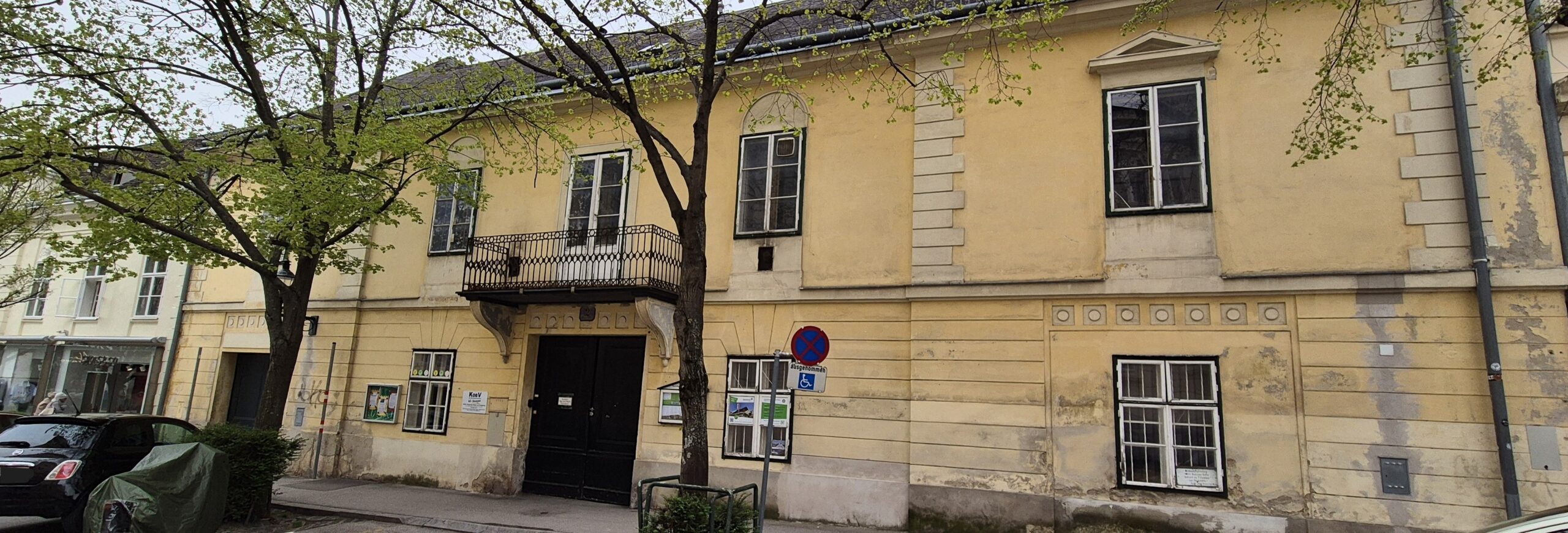 Seitlicher Blick auf ein zweigeschossiges, klassizistisches Stadthaus mit gelber Fassade und fünf Fensterachsen; davor parkende Autos.