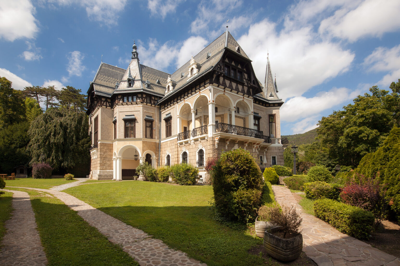 Zweigeschossige Villa mit steilen Schopfwalmdächern und reich dekorierter Ziegelfassade, davor Wiese und Wege.