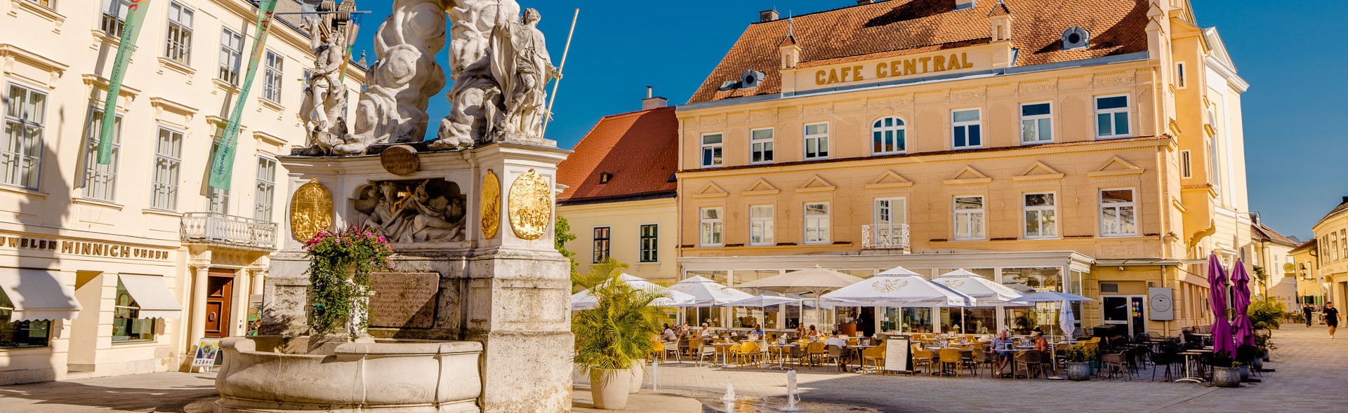 Belebter Platz mit Pestsäule; dahinter Kaffeehaus „Café Central" mit kleinem Balkon über Glasvorbau; Farbfoto.